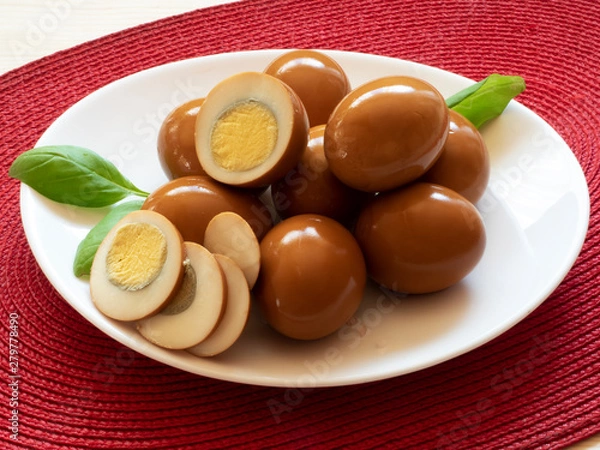 Fototapeta Brown marinated, pickled eggs, sliced egg slices.on a white plate, on a red mat, on a light table top