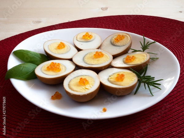 Fototapeta Pickled, marinated, egg halves decorated with caviar and greens on a white plate on a red mat, light table top