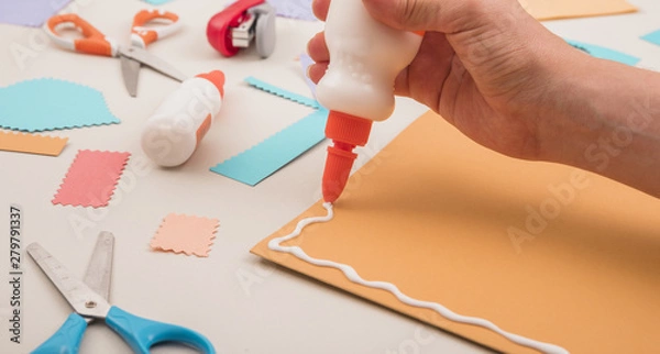 Fototapeta Human hand applying white glue on orange paper with scissor and stapler