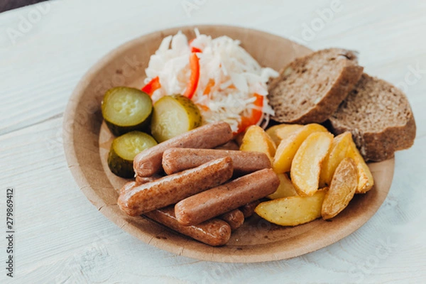 Fototapeta Tasty sausages with fried potatoes, cucumber, cabbage and chlobe in a paper plate on a wooden table.