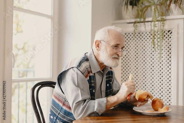 Fototapeta Lonely elderly man eating an apple by the table in empty home