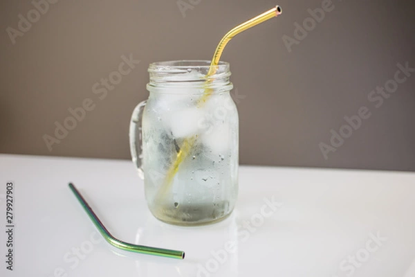 Fototapeta mason jar with ice cold water and reusable metal steel golden eco friendly straw on white table with another green bright straw on a side.