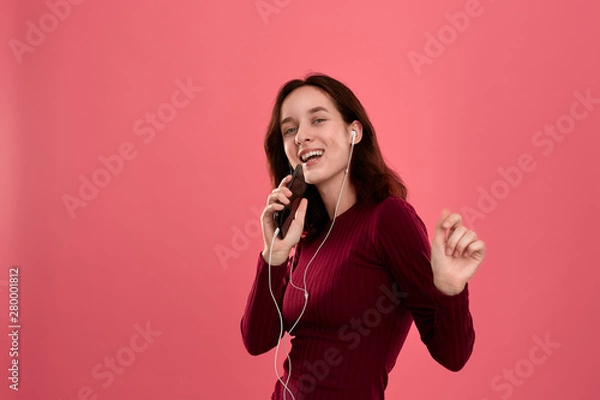 Fototapeta Happy young brunette woman singing and dancing using smartphone as a microphone and wearing wired headset.