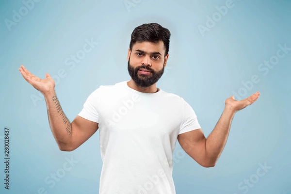 Fototapeta Half-length close up portrait of young hindoo man in white shirt on blue background. Human emotions, facial expression, ad concept. Negative space. Showing empty bar, pointing, choosing, inviting.