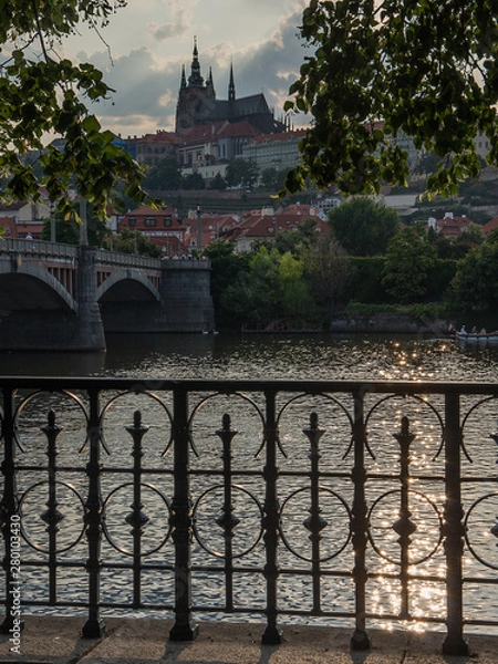Obraz Prague castle on summer evening. Back lit seen across Vltava river with a brodge, cast iron railing and sunlit green eaves in foreground