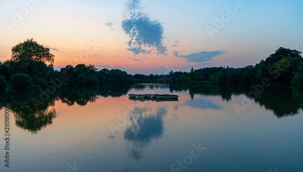 Obraz Sunset, twilight view of orange, pink and blue sky reflected in tranquill lake with rowing boats silhouette to centre of frame