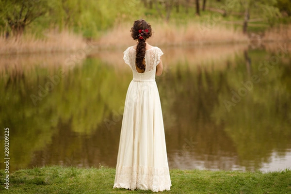 Fototapeta Rear view of a young anonymous bride in a beautiful full wedding dress, with hairstyle, looking down, nature background.