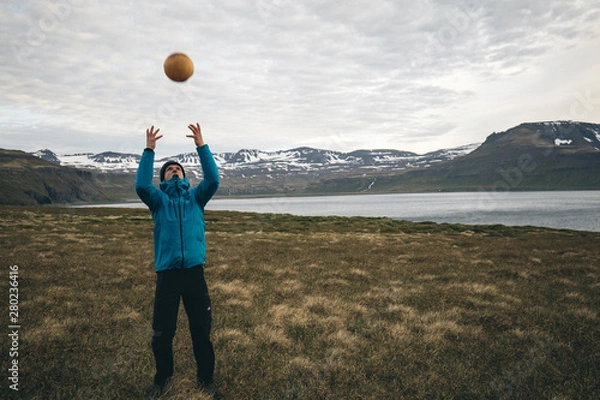 Fototapeta guy in blue jacket and black sporty pants throwing a ball up high in nature