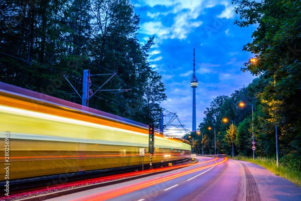 Fototapeta Germany, Illuminated tramway passing by street of traffic next to television tower of stuttgart city in green forest nature landscape in magical twilight mood after sunset