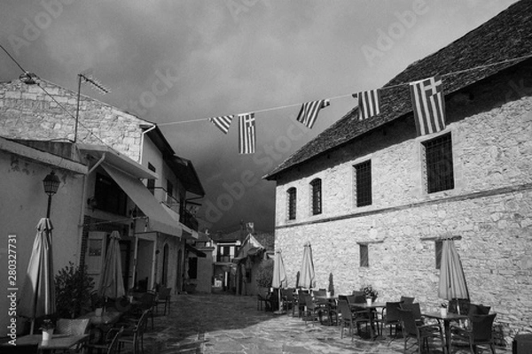 Fototapeta Perspective of street with cafe in old town. Architecture in rustic styles. White stone walls and dark dramatic sky. Tables, chairs, umbrellas and flags