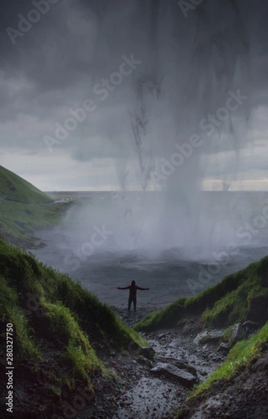Fototapeta Silhouette of a man standing under the huge waterfall. Visible Streams of water. Dramatic cloudy sky. Iceland