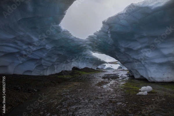 Fototapeta Small river flowing through the arch of blue snow. Ice bridge with big holes on the cloudy grey sky background. Landmannalaugar, Iceland