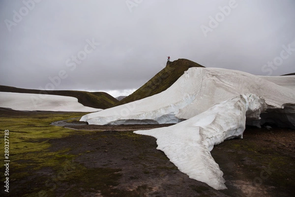 Fototapeta Small silhouette of a man on top of the mountain showing direction. Hills, green field, river and snow with cloudy background. Landmannalaugar trek, Iceland