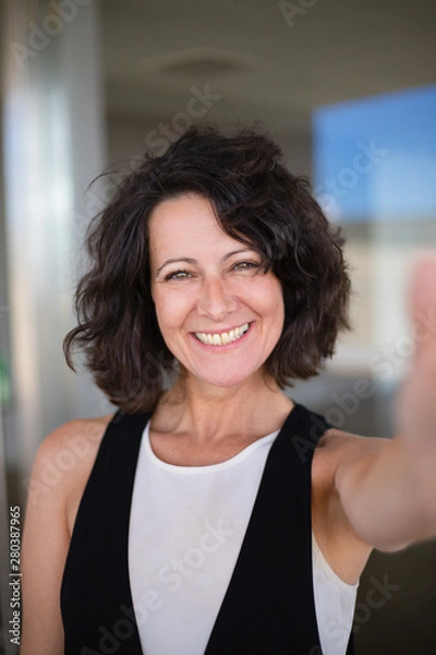 Fototapeta Selfie of happy joyful curly haired woman in casual. Excited middle aged lady holding smartphone and taking picture of herself. Happy woman portrait concept