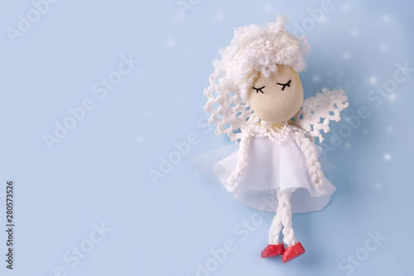 Fototapeta Postcard layout with a cute handmade angel in white clothes on a light blue background with bokeh. Copy space