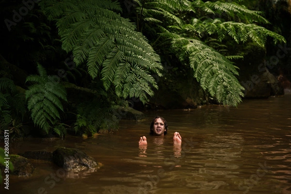 Fototapeta A curly man swimming on his back on the silty brown water with face and feet raised up. Tropical river covered with big fern leaves