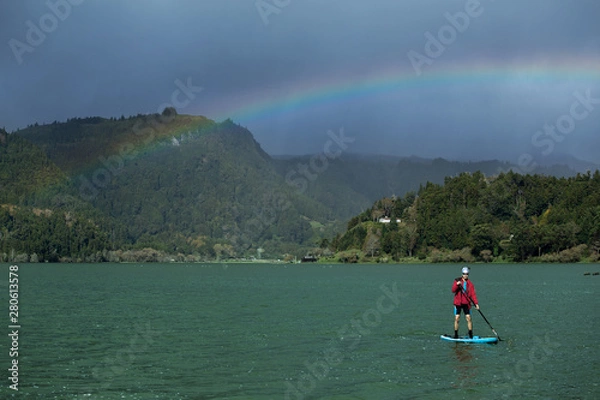 Fototapeta A man in shorts and red jacket floats standing on a sapsurf on the lake in cloudy weather with beautiful rainbow background. Furnas Lake, Azores
