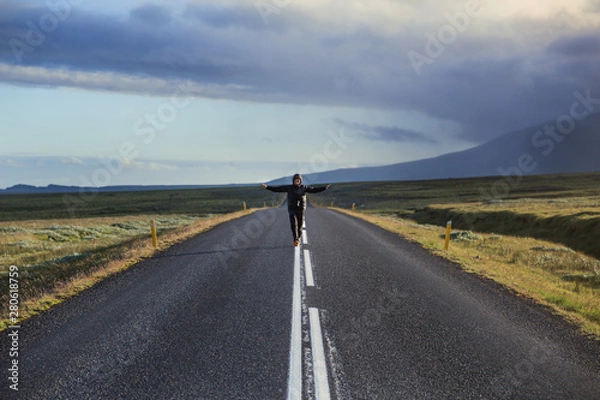 Fototapeta Man in black clothes is moving towards right in the middle of the long road through fields with mountain background. Side sunset light. Iceland