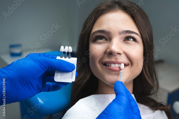 Fototapeta dentist in blue medical gloves applying sample from tooth enamel scale to happy woman patient teeth to pick up right shade for teeth bleaching procedure