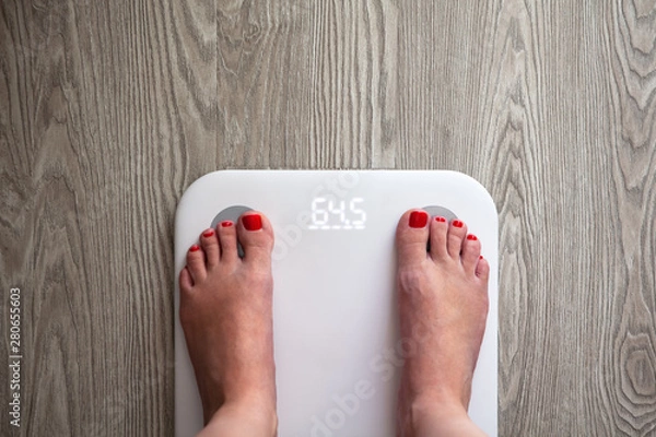 Fototapeta Woman stands on white modern electronic scales, which show 64.5 kg. Only feet are visible. Scales stand on gray wooden floor. Space for text. Healthy lifestyle, diet, weight loss concept. Horizontal.