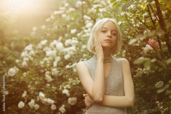 Fototapeta Portrait of beautiful young girl with hands on her face in white roses. Blond platinum girl with fair complexion. Sunset backlight. Image with selective focus and toning