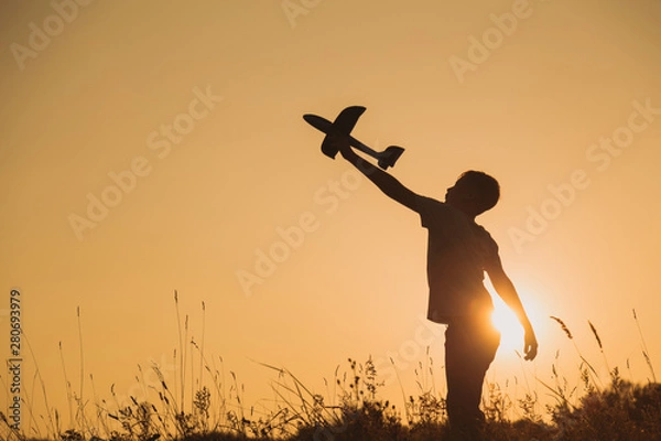 Obraz Black silhouette of young caucasian kid isolated on sunny golden sunset sky background. Boy playing toy plane outside on grassy summer hill. Setting big goals and dreaming about happy future concept.
