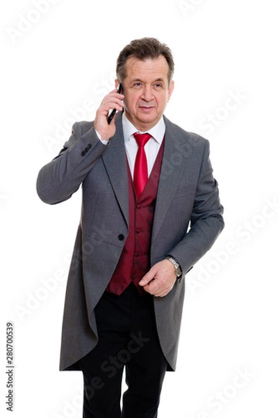 Fototapeta Portrait of handsome confident caucasian mature businessman 55-60 years old in grey tuxedo and red vest. Rich senior man talk by phone and looking to camera on isolated white background