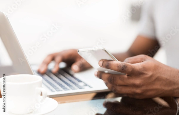 Fototapeta Unrecognizable african man using cellphone and laptop