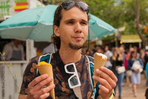 Fototapeta Close up portrait of a handsome young man savouring two ice cream cones over fair background. Photographer with sunglasses and backpack.