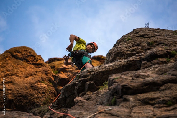 Fototapeta Low angle view of a man climbing on cliff against blue sky. Rope, helmet and all equipment.