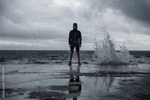 Fototapeta Portrait of fit man on the pier in shorts and waterproof jacket with a hood. Dramatic background with beating wave and reflection on the floor. Feeling of freedom. Sao Miguel island, Azores.