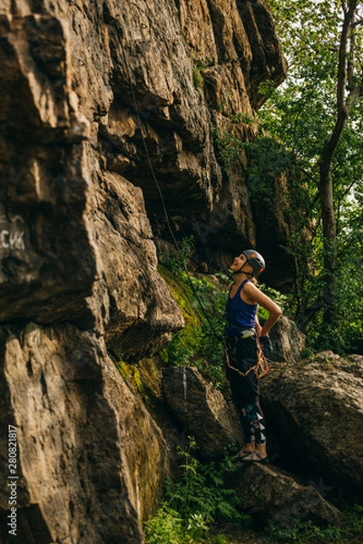 Fototapeta Young beautiful girl wearing in climbing equipment standing in front of a stone rock and preparing to climb against brown cracked rock and trees