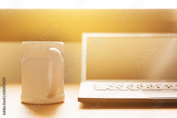 Fototapeta laptop is on the desk at home near window in the morning. there is a cup of coffee for refresh you from awake. the sun is shining from outside onto the desk.