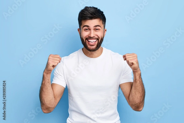 Fototapeta excited Arab man celebrating success with two fists in air isolated on the blue background. close up portrait, studio shot , happiness, positive emotion and feeling. I've done it. facial expression