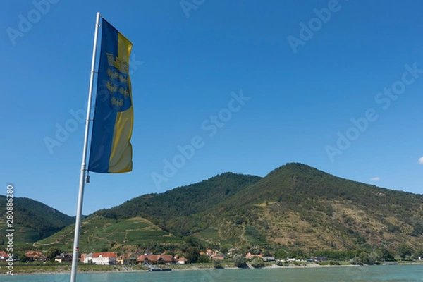 Fototapeta Flag of the country Lower Austria in the Wachau in front of the Danube with a mountain with vineyards in the background