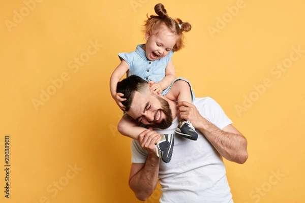 Fototapeta little lovely girl tickling her daddy while sitting on his shoulders . isolated yellow background, studio shot.feeling and emotion