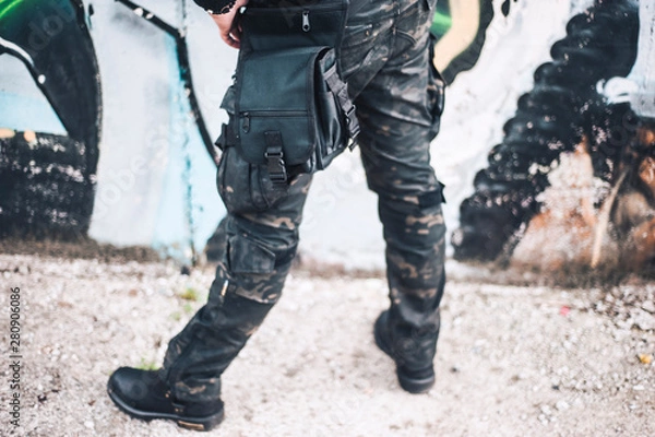 Fototapeta An urban man in a camouflage military pants stands next to the wall. Background from the urban part of the city