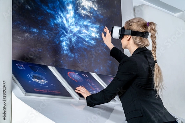 Fototapeta A girl in a black jacket standing in front of control panel monitors in virtual reality glasses. Looking forward, holds hands in front of the panels. Middle shot. Spaceship interior. Low angle
