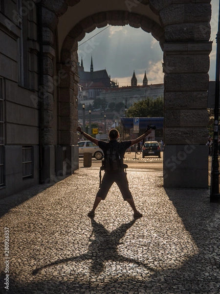 Obraz Fun in Prague - silhouette of a man posing beneath an arcade. He is turning his back to camera and throwing his shadow on a sidewalk in a stark back light. Sun lit Prague castle in distance.