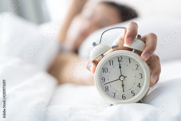 Fototapeta Closeup woman sleeping on bed and hand holding alarm on clock, Time to wake up