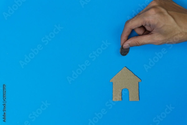 Fototapeta Property investment, savings money for buy new home concepts. A man hand holding coin over small residence house in paper on blue background and space. A sustainable investment.
