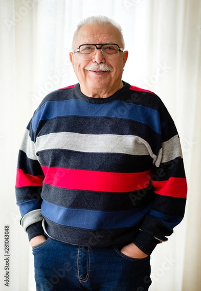 Fototapeta Portrait of smiling and confident senior man 70-75 years old with eyeglasses stands near light window. Holding hands in pockets jeans