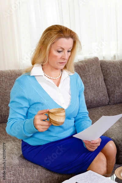Fototapeta Business middle-age woman 60-65 years old sit on grey sofa and works at home. Senior female reads the letter with a surprised expression with cup in hand.