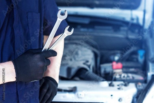Fototapeta Repairman is repairing car at service station. Closeup mechanic hands in gloves are holding steel wrenches. Vehicle with open hood on background.  Modern auto repair shop with equipments and  tools.