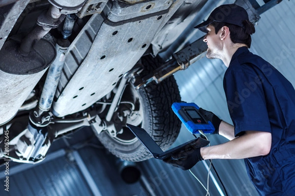 Fototapeta Mechanic is repairing car at service station. Repairman is detecting problems by special software on laptop and diagnostic tool at workshop auto repair shop. Vehicle on hydraulic lift is above.