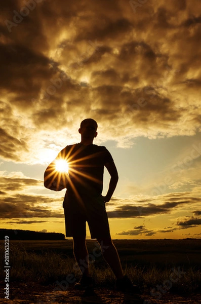 Fototapeta Silhouette of a man with a short haircut in a T-shirt and shorts with a ball. The concept of games with a ball in the fresh air, volleyball. Photo in warm sunset colors. Place for text. 