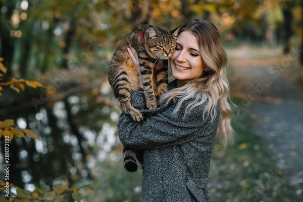 Fototapeta girl and a cat in the autumn park.a woman in a brown sweater walking with her gray cat in an autumn park