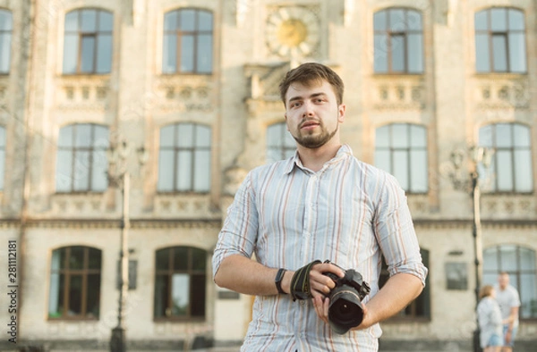 Obraz Portrait of a young handsome bearded man in striped shirt holding professional camera, shooting outdoors, stylishly dressed, photographer, close up, brutal, street photo, architecture background.