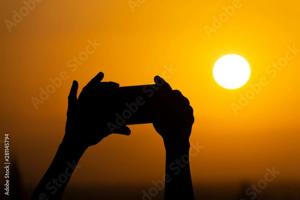 Fototapeta Silhouette of hands holding a mobile phone and making photography of the huge sun during sunset or sunrise. Male makes a photo on his phone.