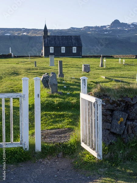 Obraz Black Church in Budir - PoI in western Iceland. The wooden church seen in late evening sun illumination through an open white painted wooden gate across an old cemetery. Mountains in background